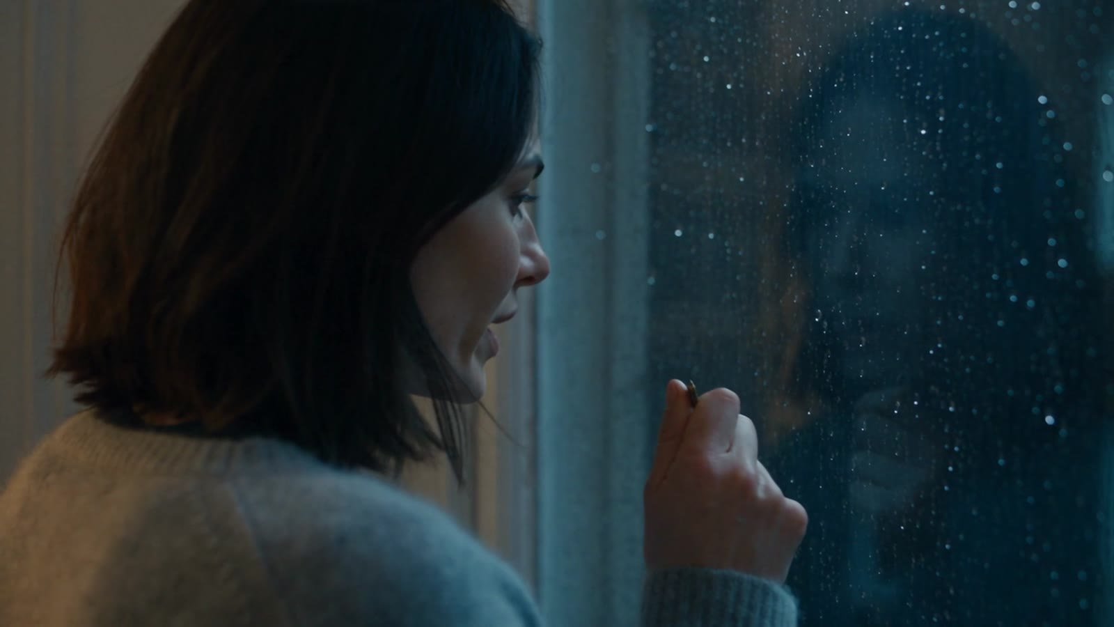 A woman by a rainy window holding a small key while a man looks at her, in a dim apartment