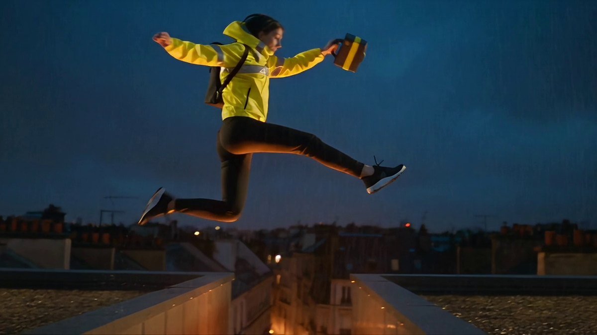 A courier in a safety-yellow shell jacket mid-leap between two wet rooftops at night